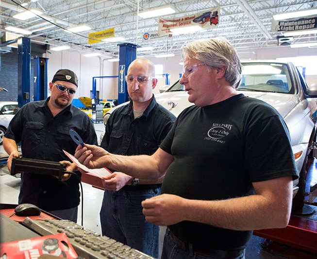 three men in a workshop examining parts of a car